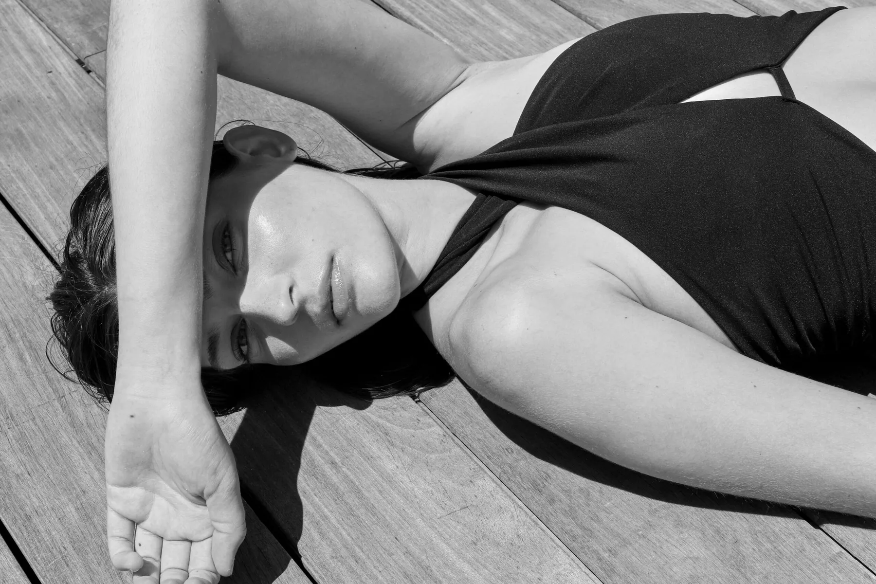 Black and white: model in a black swimsuit lying on a wooden deck, arm over her eyes