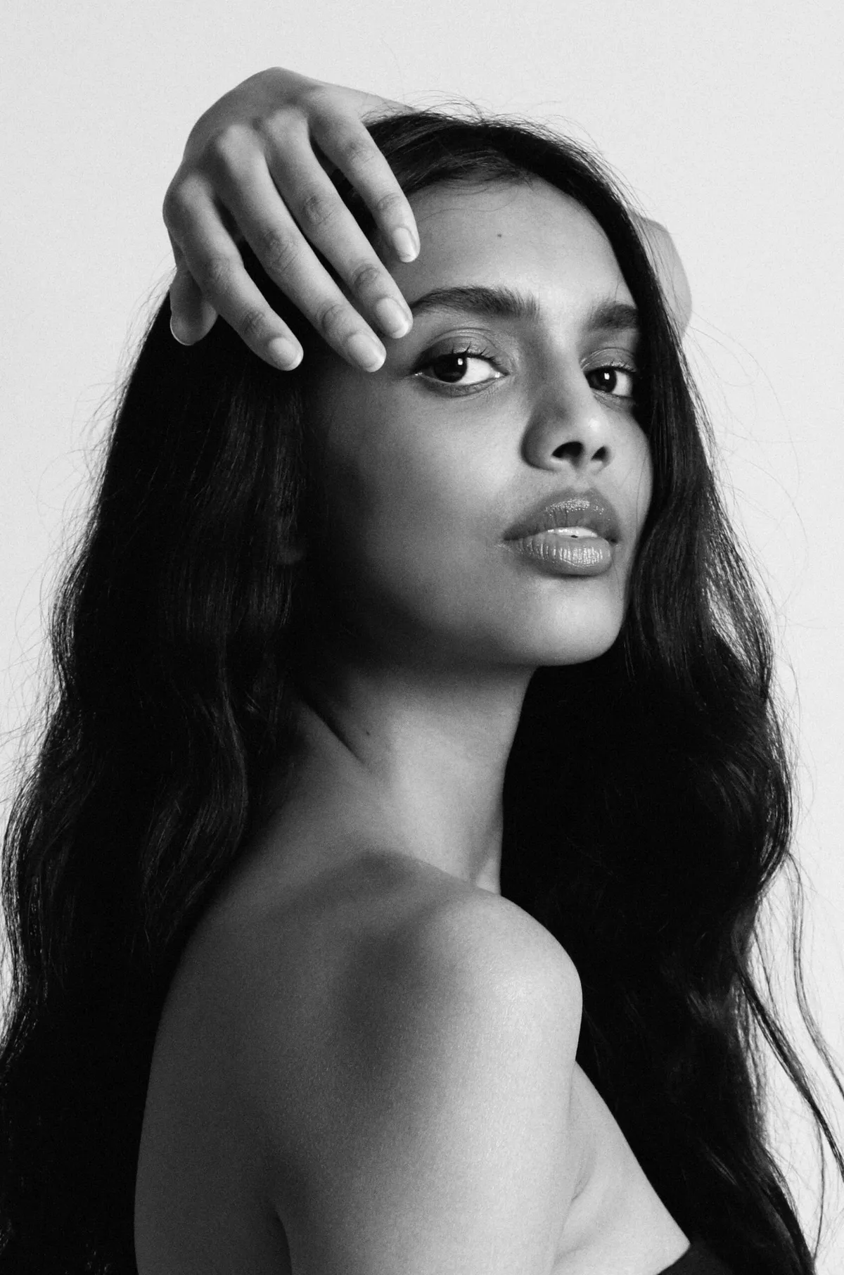 Black and white portrait: model with long dark hair, one hand resting on her forehead, looking past the camera