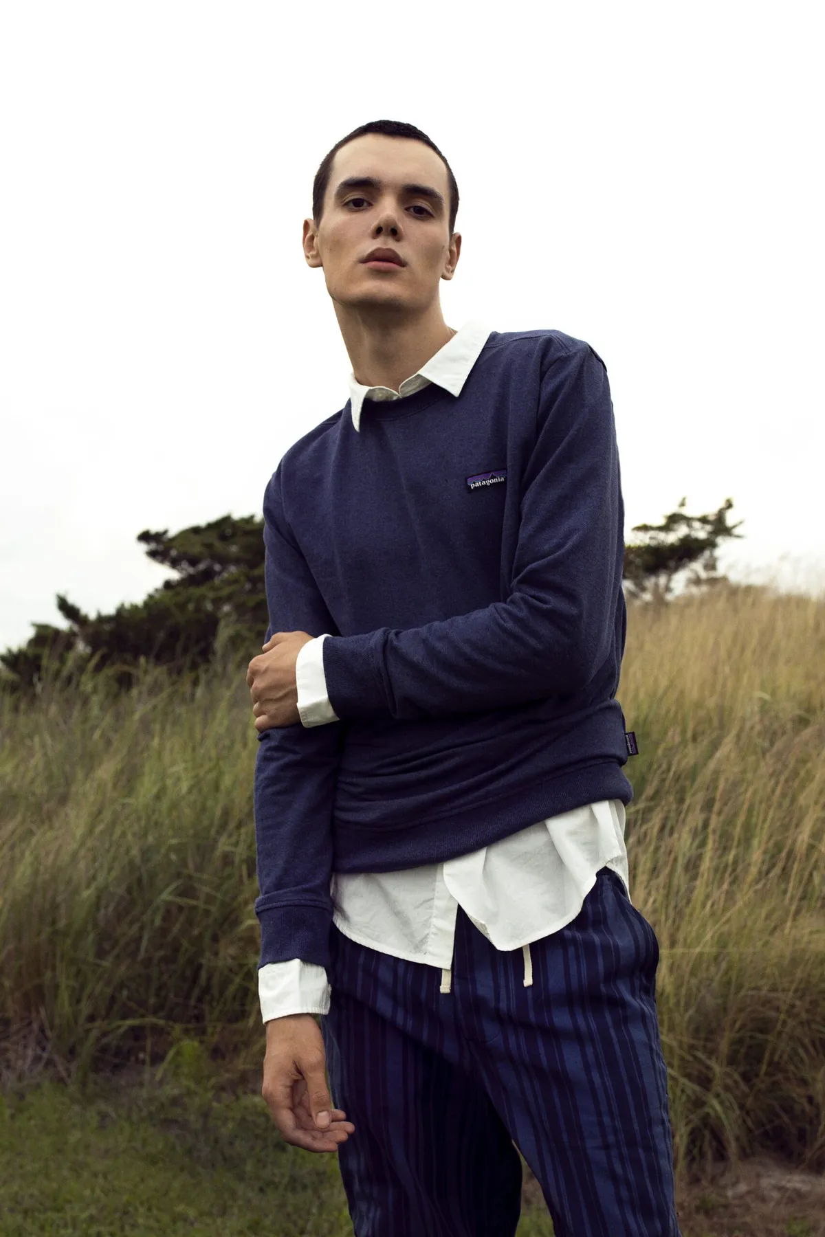 Man in a navy polo over a white shirt and striped trousers, buttoning a cuff, dune grass behind