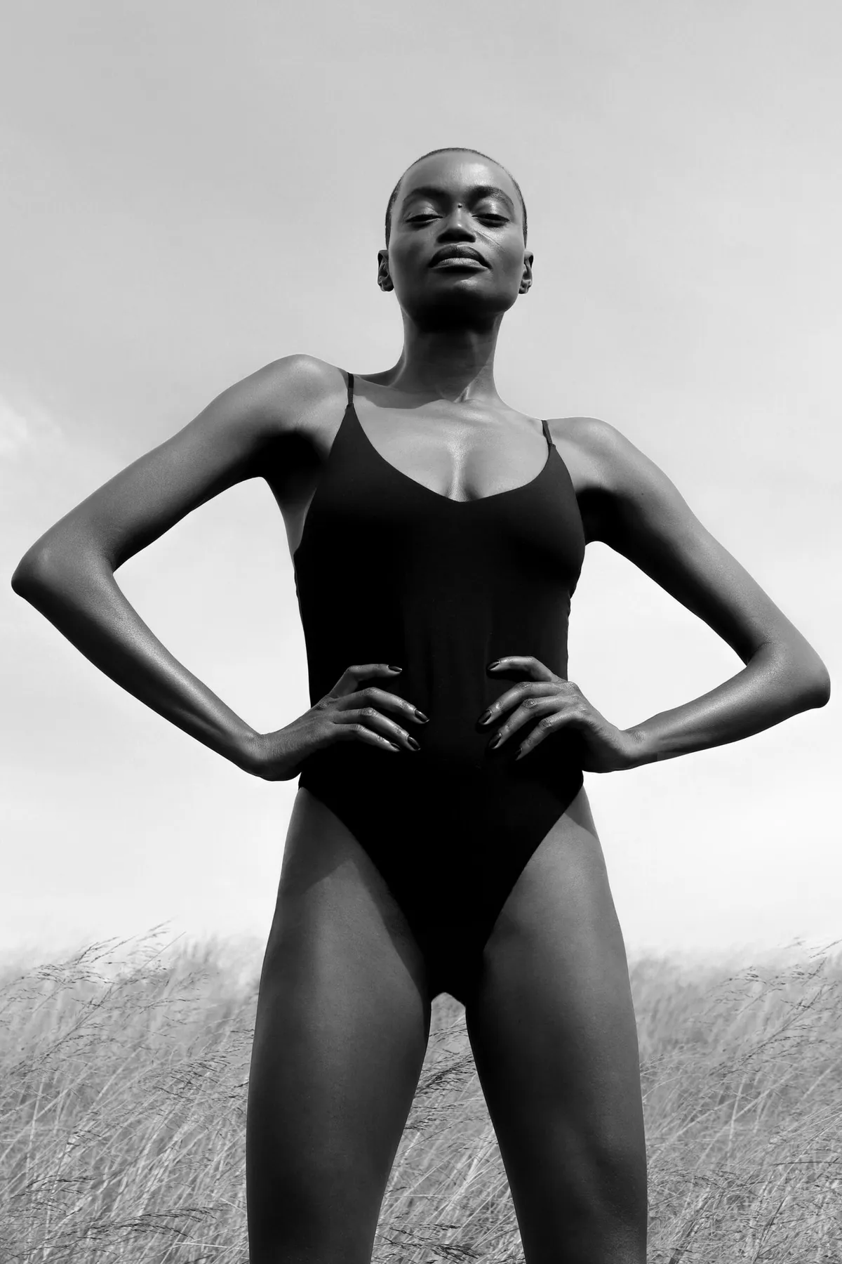 Black and white: model in a black one-piece swimsuit, hands on hips, against an overcast sky