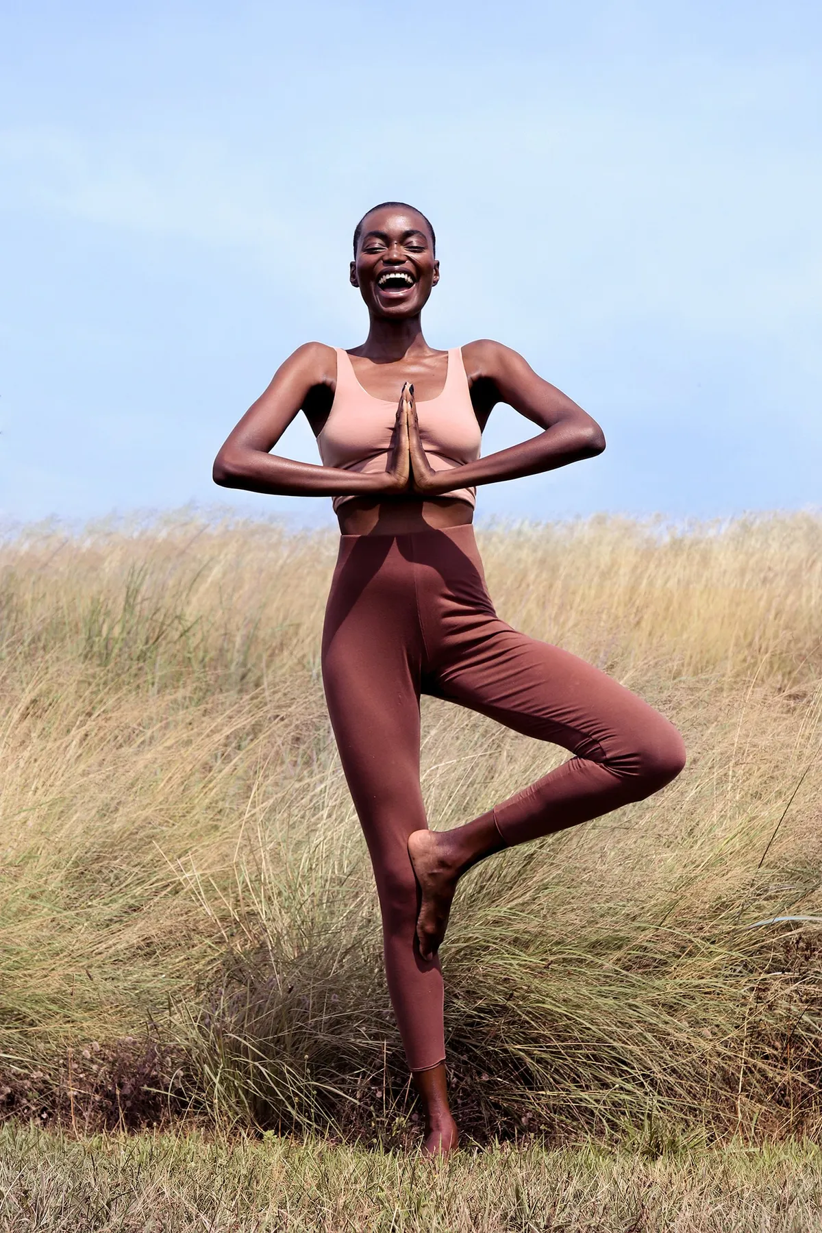 Model in a rust-coloured top and leggings balancing in tree pose in a golden field
