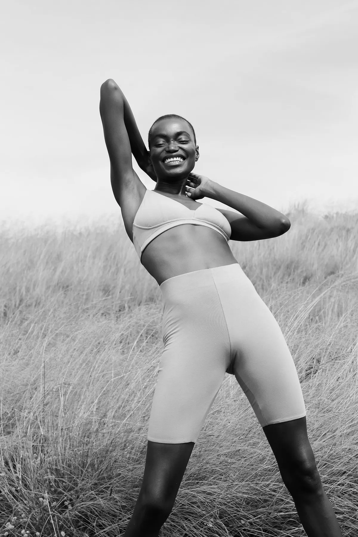 Model in a pale bralette and bike shorts laughing with one hand behind her head, black and white