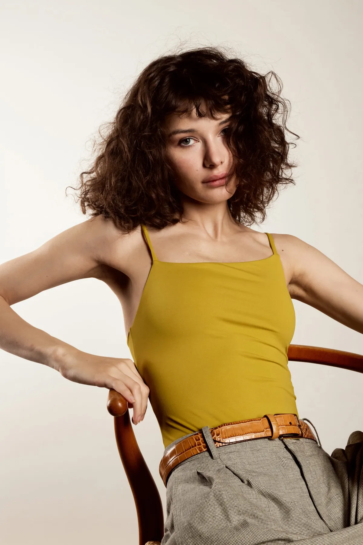 Model in a mustard camisole and belted grey trousers seated on a wooden chair, looking to camera