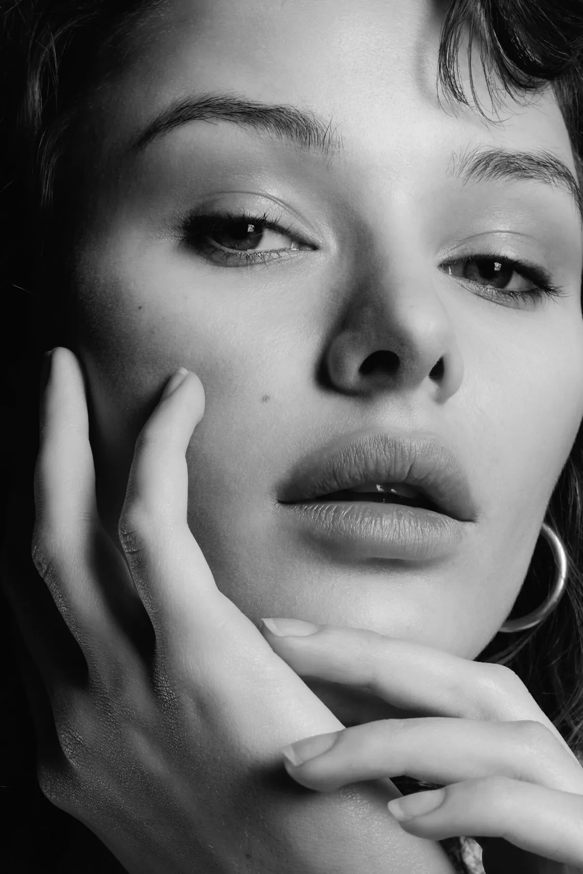 Black and white close-up: a model’s face half covered by her hands, a hoop earring