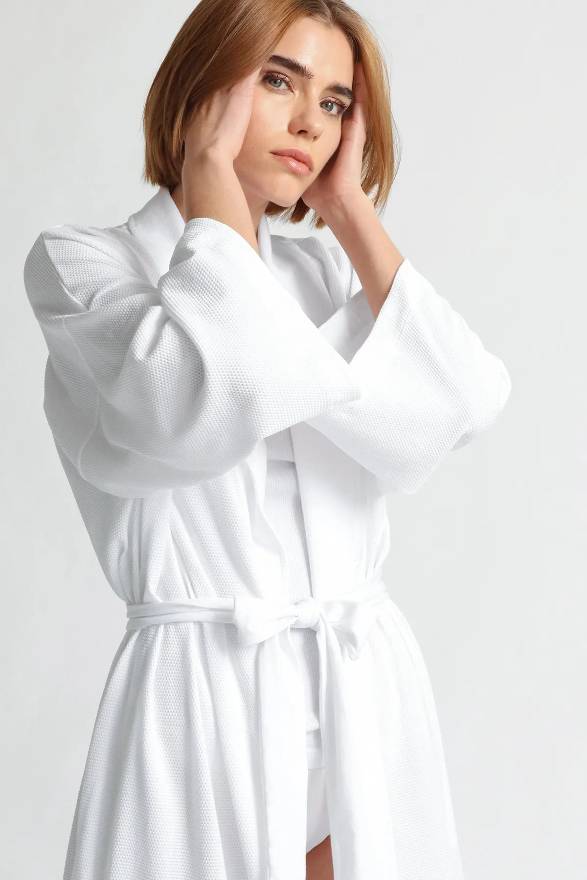 Model in a white wrap robe, hands lifted to her temples, looking to camera